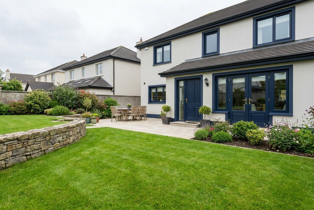 A freshly levelled Irish back garden with a flat striped lawn, a low natural-stone retaining wall, a pale-stone patio, and navy French doors with brass hardware on the house rear under overcast Irish daylight