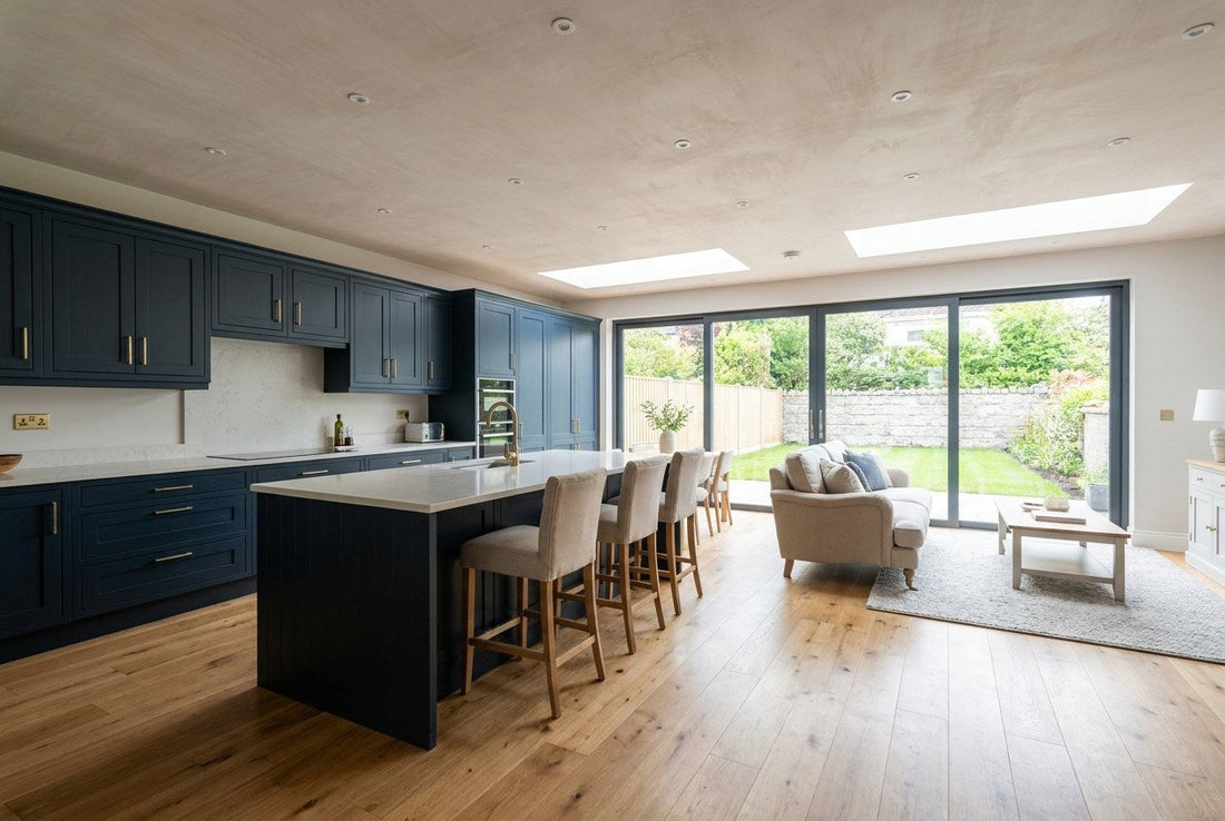 A finished open-plan kitchen and living space in a renovated Irish home, with deep navy shaker cabinetry, brass handles, a pale stone worktop and glazed doors onto the garden